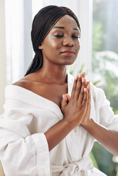 Κάθετο πορτρέτο της όμορφης Αφρικανής γυναίκας που φοράει λευκό μπουρνούζι και διαλογίζεται στο σπίτι με τα χέρια της προσευχής - Φωτογραφία, εικόνα