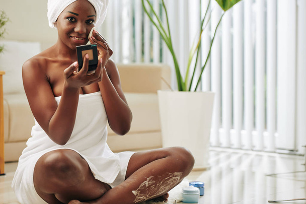 Retrato horizontal de atraente jovem afro-americana usando toalha de banho fazendo sua rotina diária de cuidados com a pele
 - Foto, Imagem
