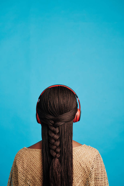 Vertikální zadní pohled záběr nerozpoznatelné černošky žena s dlouhými vlasy nosí červené sluchátka poslech hudby, modré pozadí - Fotografie, Obrázek