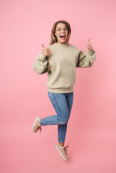 Portrait of young excited woman in eyeglasses jumping with thumbs up - Foto, imagen