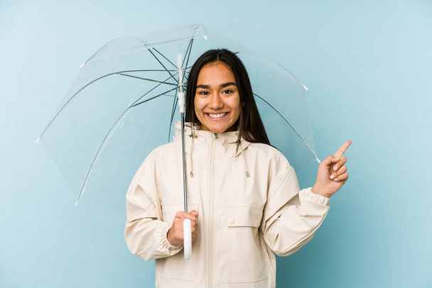 Joven asiática sosteniendo un paraguas sonriendo y señalando a un lado, mostrando algo en espacio en blanco
. - Foto, Imagen