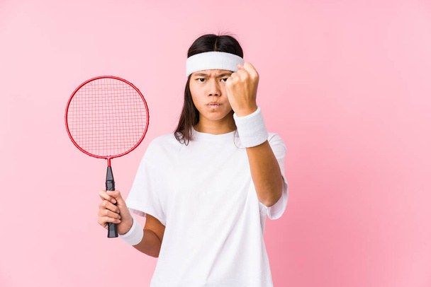 Joven mujer china jugando bádminton en un fondo rosa mostrando puño a cámara, expresión facial agresiva
. - Foto, imagen
