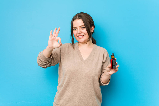 Jovem mulher curvilínea segurando um vaporizador alegre e confiante mostrando gesto ok
. - Foto, Imagem