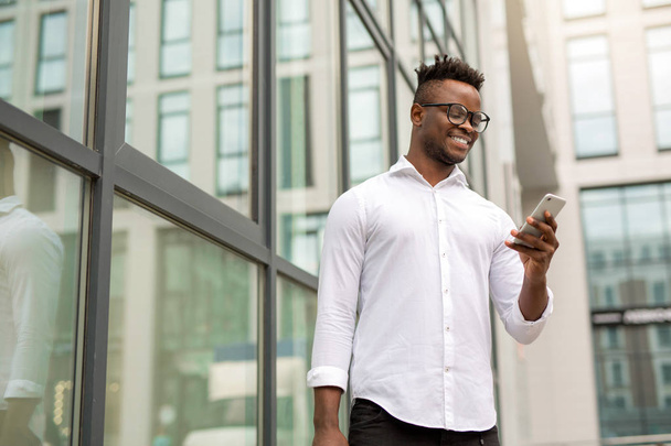 handsome young african man in a white shirt with a phone in his hands near a glass building - Foto, Imagem