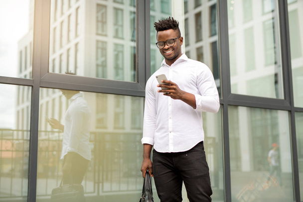 handsome young african man in a white shirt with a phone in his hands near a glass building - Foto, immagini