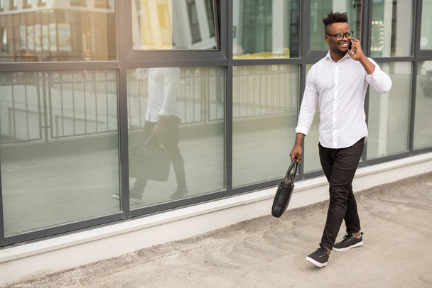 handsome young african man in a white shirt with a phone in his hands near a glass building - Фото, изображение
