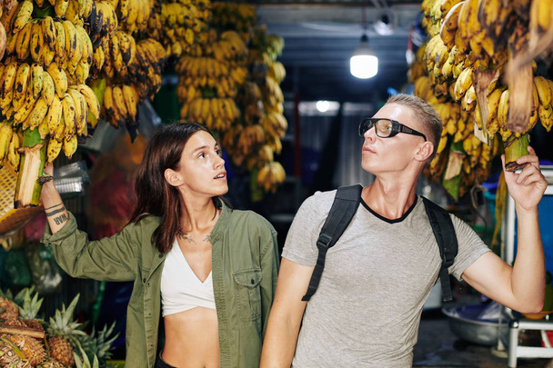 Young man and woman walking along Asian farmers market looking at bananas and chatting, horizontal shot - Photo, Image