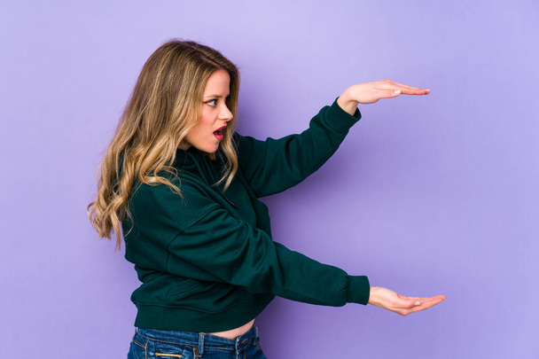 Young caucasian woman isolated on purple background shocked and amazed holding a copy space between hands. - Foto, Imagen