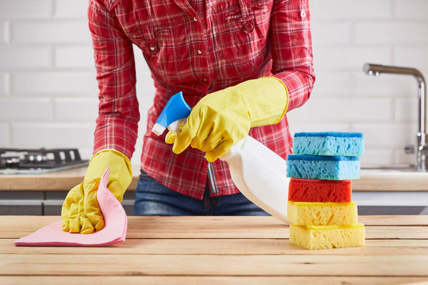 woman in yellow rubber protective gloves cleaning wooden table with spray, sponge and cloth, kitchen background - Фото, зображення