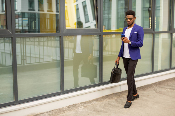Όμορφος νεαρός Αφρικανός με κοστούμι και τηλέφωνο κοντά σε γυάλινο κτίριο. - Φωτογραφία, εικόνα
