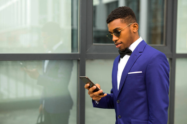 handsome young african man in a suit with a phone in his hands near a glass building - Foto, afbeelding