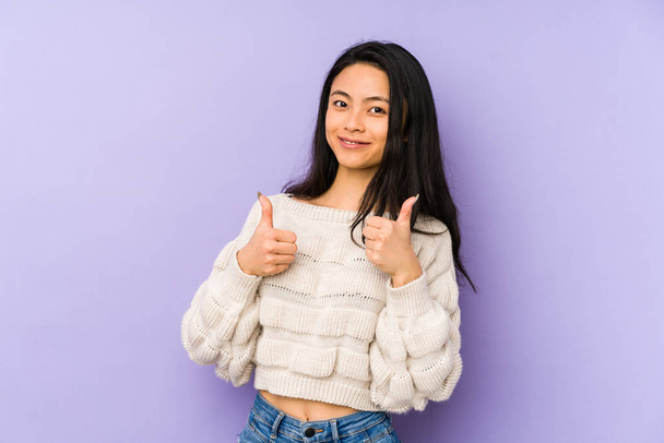 Jonge Chinese vrouw geïsoleerd op een paarse achtergrond het verhogen van beide duimen omhoog, glimlachen en zelfverzekerd. - Foto, afbeelding