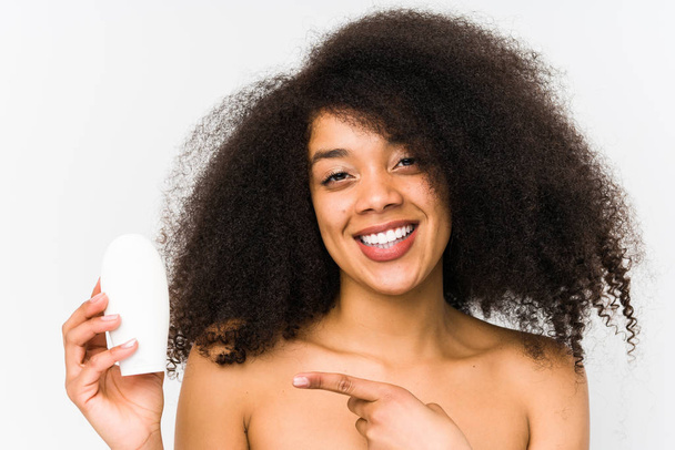 Junge Afro-Frau hält isoliert eine Feuchtigkeitscreme und lächelt fröhlich und zeigt mit dem Zeigefinger weg. - Foto, Bild