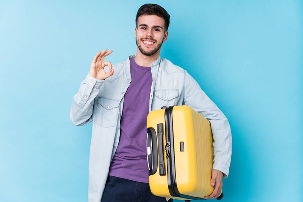 Jeune caucasien voyageur homme tenant une valise isolé gai et confiant montrant ok geste
. - Photo, image