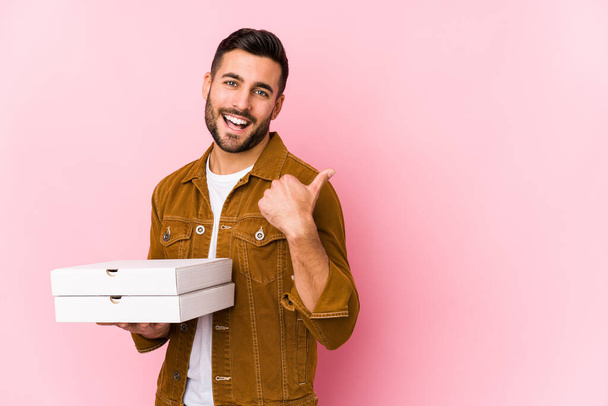 Joven hombre guapo sosteniendo pizzas puntos aislados con el dedo pulgar lejos, riendo y sin preocupaciones
. - Foto, imagen