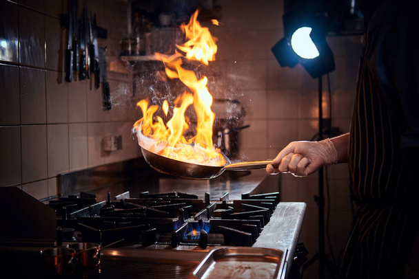 Fogão usando luvas e avental de fritura flambe em uma panela em uma cozinha restaurante escuro
 - Foto, Imagem