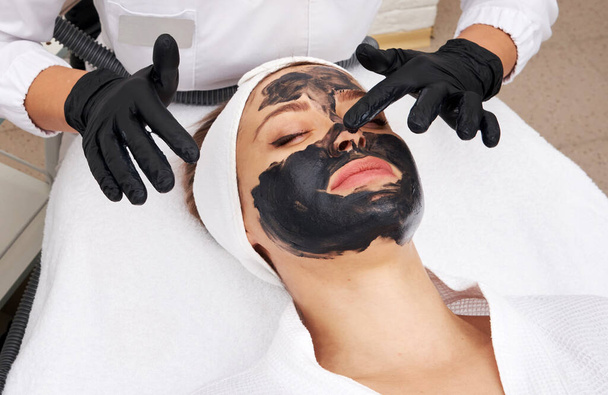 Kozmetológus fekete maszkot visel egy gyönyörű nő arcán szénhámlásra. - Fotó, kép