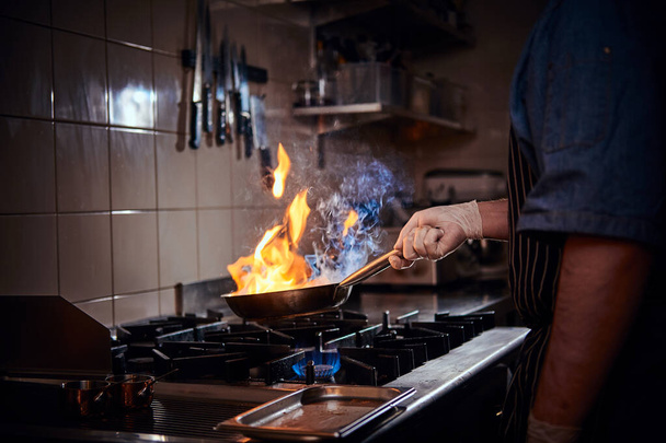 Vařič v rukavicích a zástěře smažení flambe na pánvi v tmavé restauraci kuchyně - Fotografie, Obrázek