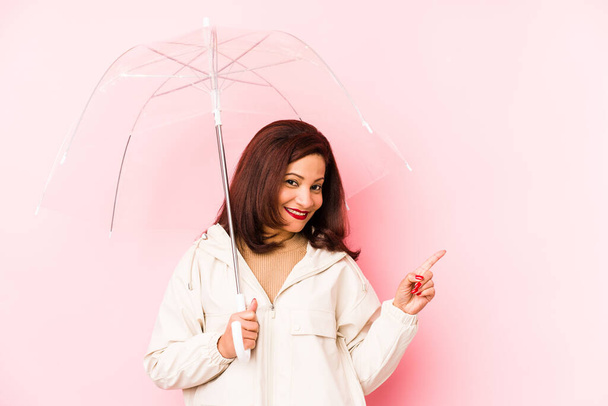 Lateinische Frau mittleren Alters, die einen Regenschirm trägt, isoliert lächelnd zur Seite zeigt und etwas im leeren Raum zeigt. - Foto, Bild