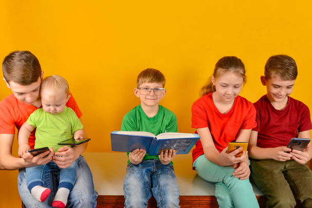 子供たちは隣に座って電話を見ているが、眼鏡をかけた少年は本を持って座っている。家庭でのオンライン学習の概念. - 写真・画像