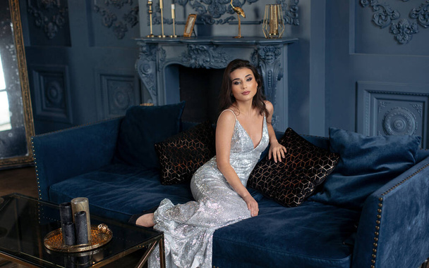 Эффективная брюнетка в серебристом блестящем платье позирует на диване в шикарном винтажном интерьере
 - Фото, изображение