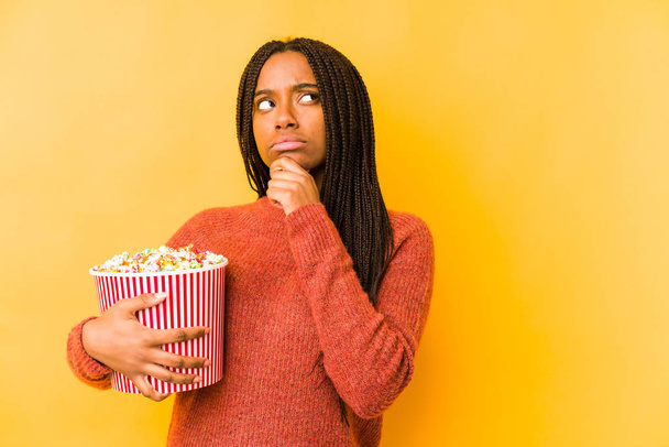 Young african american woman holding a popcorn isolated looking sideways with doubtful and skeptical expression. - Photo, Image