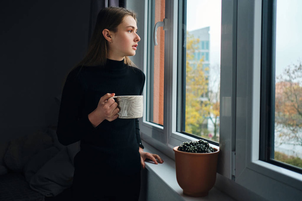 Side view of attractive girl holding mug thoughtfully looking out the window at home  - Photo, Image