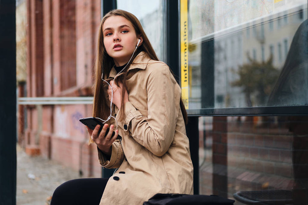 Menina casual atraente em fones de ouvido com celular esperando atenciosamente transporte público na parada de ônibus ao ar livre
 - Foto, Imagem