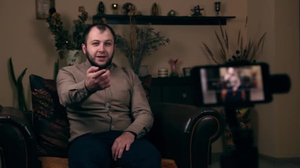 Wpływowy Brodaty Młody, bloger o europejskim wyglądzie, siedzi na krześle w koszuli, nagrywa wideo przez telefon i prosi o komentarz pod filmem. Koncepcja blogowania mediów społecznościowych - Materiał filmowy, wideo