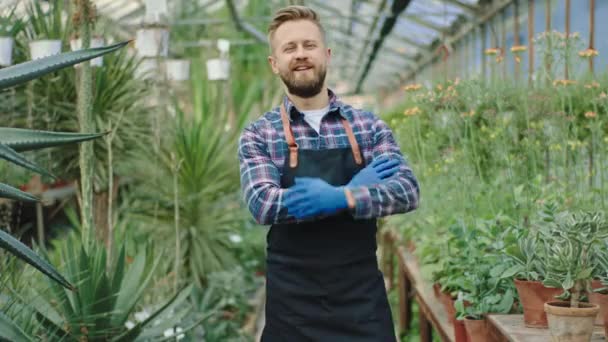Happy gar portrait in front of the camera he crossed hands and smiling charismatic looking straight to the camera background a very beautiful flower greenhouse - Video