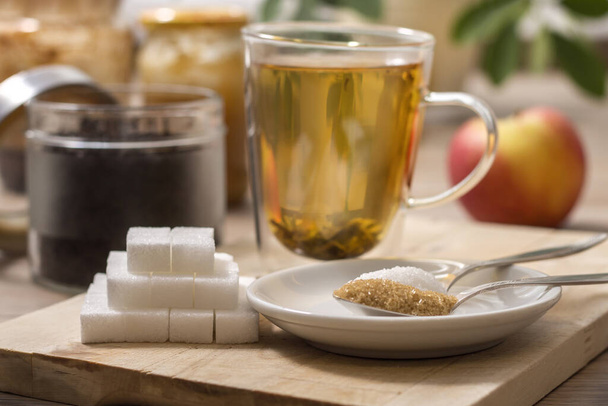 Klumpen Zucker, Rohrzucker, loser weißer Zucker und Buchweizenhonig liegen auf einem Teelöffel neben einem Glas mit aufgebrühtem Kräutertee.. - Foto, Bild
