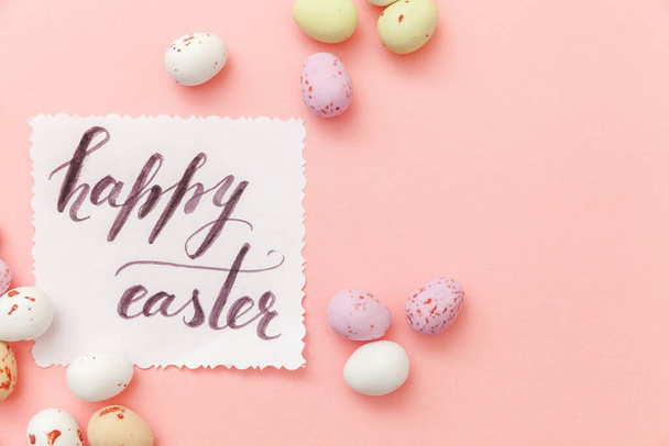 Happy Easter concept. Inscription HAPPY EASTER letters candy chocolate eggs and jellybean sweets isolated on trendy pastel pink background. Simple minimalism flat lay top view copy space. - Photo, image