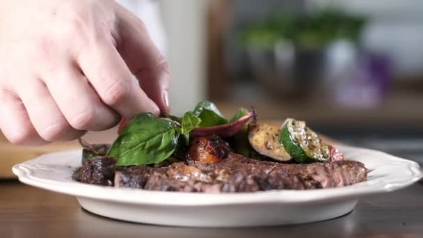 a szakács friss bazsalikomot tesz egy tányérra, grillezett marhahúsos szűzpecsenyével és grillezett zöldségekkel. Élelmiszerdekoráció - Felvétel, videó