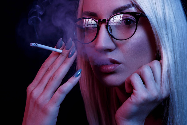 jonge blanke mooie blonde vrouw met bril roken. close-up portret in neon licht - Foto, afbeelding