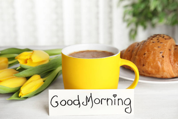 Croissant, kawa, piękne kwiaty i kartka ze słowami Dzień dobry na białym drewnianym stole wewnątrz - Zdjęcie, obraz