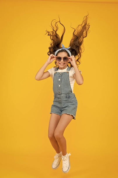 Set a truly energetic and optimistic mood. Energetic little girl with long brunette hair jumping on yellow background. Active small child with cute look feel energetic. Energetic fashion style - Fotografie, Obrázek
