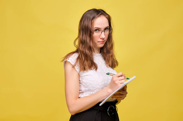 Tiukka opettaja tyttö, jolla on lasit keltaisella taustalla, tekee muistiinpanoja kynällä muistikirjassa.
. - Valokuva, kuva