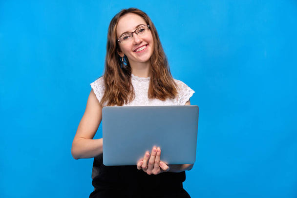 A brunette girl in casual clothes is holding a laptop on a blue background with glasses holding a laptop and looking at the camera. - Fotó, kép