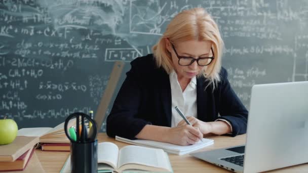 Mature woman in suit working in class writing sitting at desk with books and laptop - Footage, Video