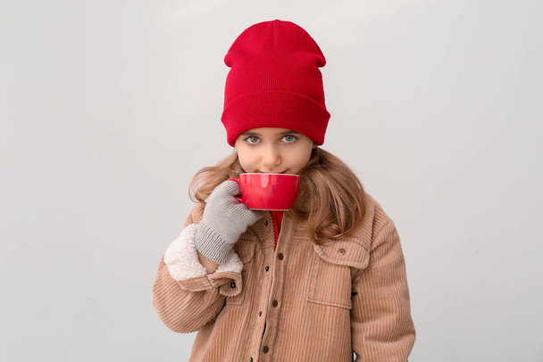 Cute little girl in winter clothes and with cup of hot chocolate on light background - Photo, Image