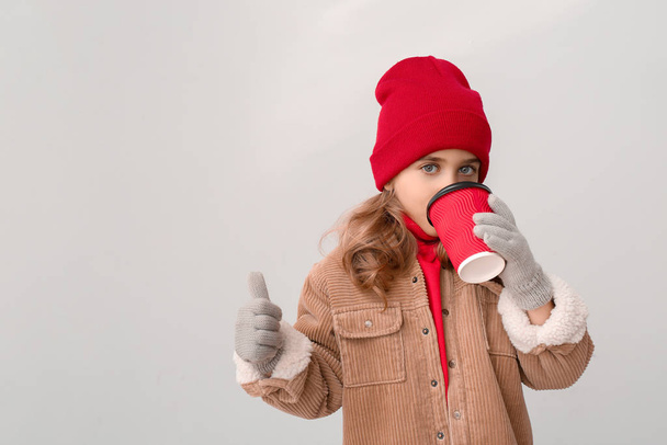 Cute little girl in winter clothes and with cup of hot chocolate showing thumb-up gesture on light background - Photo, Image