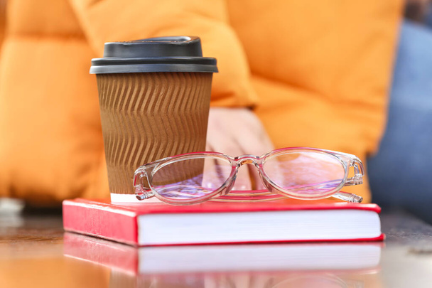 Νεαρή γυναίκα με βιβλίο, φλιτζάνι καφέ και γυαλιά που κάθεται στον πάγκο - Φωτογραφία, εικόνα