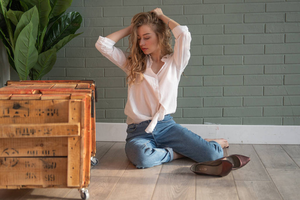 Young beautiful woman sitting on the floor with bare feet next to wooden box and green plant - Fotografie, Obrázek
