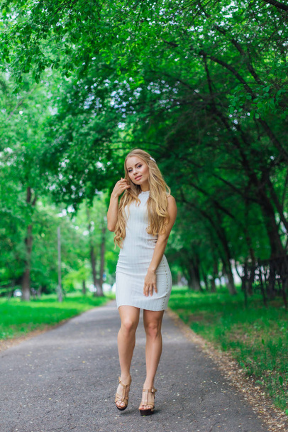 Portrait of a charming blond woman wearing beautiful white dress standing on the road under trees. - Foto, imagen