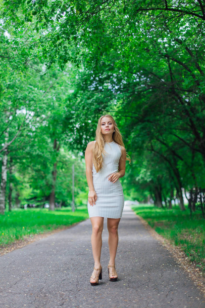 Portrait of a charming blond woman wearing beautiful white dress standing on the road under trees. - Foto, immagini