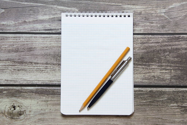 Notepad with a blank white sheet in a checker paper with ball pen and simple graphite pencil lies on the background of wooden boards  - Foto, Bild