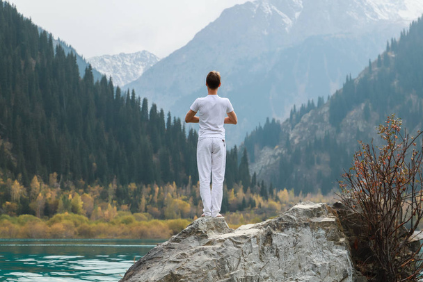 Een man staat op een steen in het midden van een bergmeer en beoefent yoga. Pose Vrikshasana. - Foto, afbeelding