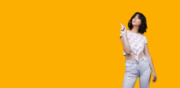 A nice view of a charming caucasian lady eating and stretching a gum on a yellow wall with freespace - Foto, Imagen