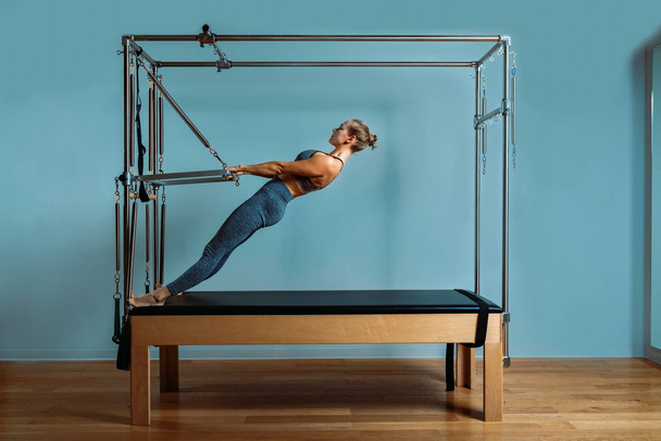 A young girl does Pilates exercises with a bed reformer, barrel machine tool. Beautiful slim fitness trainer on the background of a reformer doing various exercises gray, low key, light art. Fitness - Fotografie, Obrázek