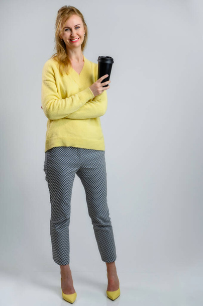 Full-length photo with a cup of coffee in the hands of a happy slender blonde woman in a yellow sweater and gray pants on a white background - Фото, изображение